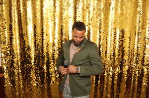 Man in green blazer holding microphone against gold sequin backdrop at glamorous event.