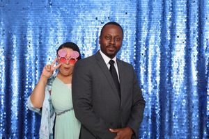 Couple posing in a photo booth with blue sequin backdrop, woman wearing oversized pink heart sunglasses.
