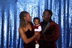 A family of three poses in front of a blue sequin backdrop, with parents holding their baby dressed in holiday attire.