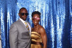 A couple smiles together in front of a blue sequin backdrop, dressed in elegant gold and gray formal attire.