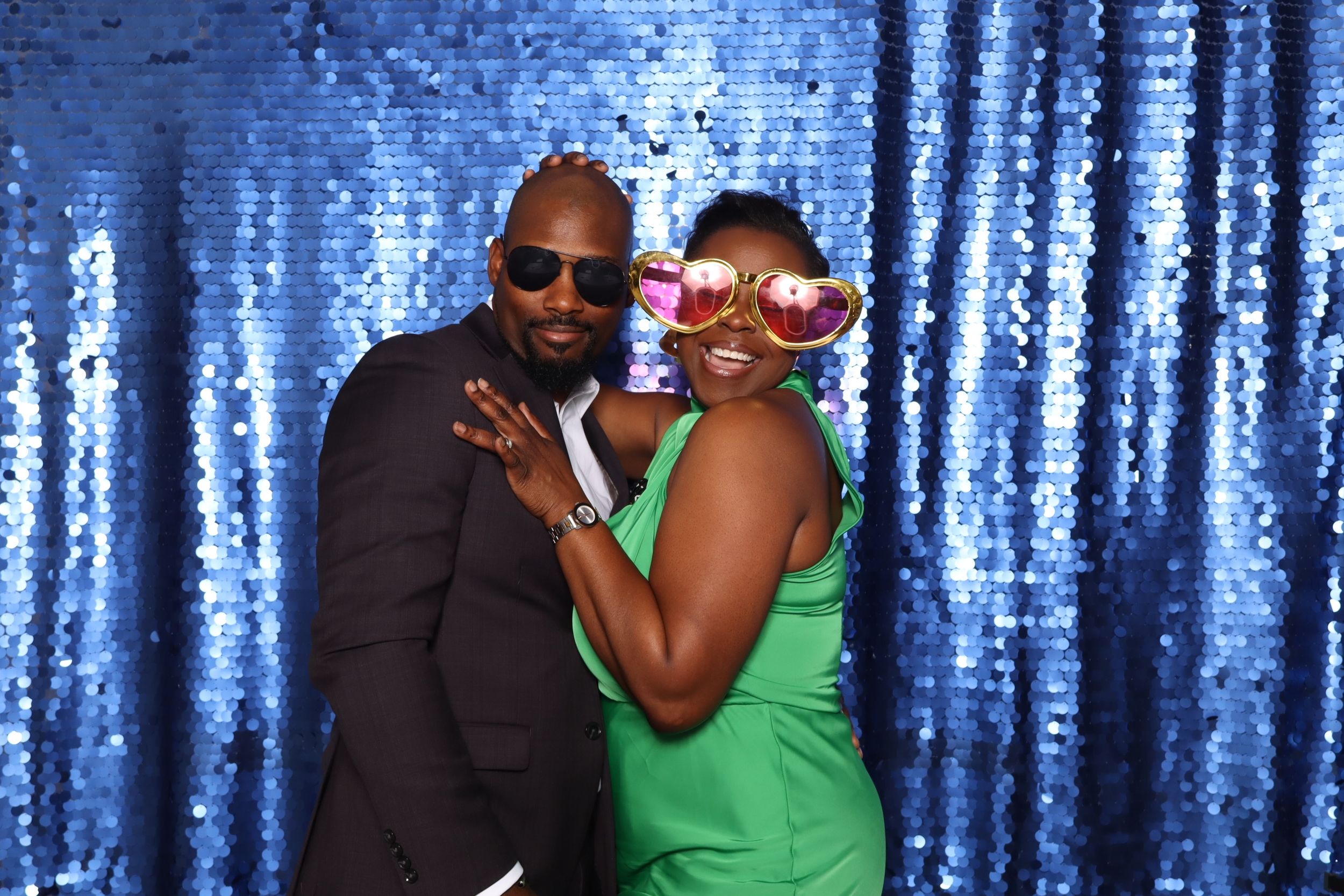 Two people posing in front of a blue sequin backdrop, one wearing fun oversized pink glasses at a party photo booth.