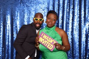 Man in suit with yellow glasses and woman in green dress pose with humorous photo booth props at party.