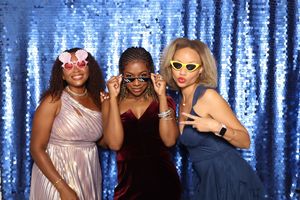 Three women posing in a photo booth with sequin backdrop, wearing colorful sunglasses and elegant dresses.