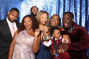 A group of seven friends pose and laugh together in front of a blue sequin backdrop at a formal event.