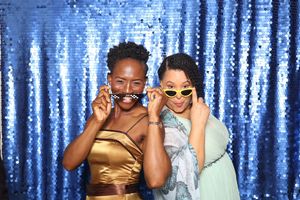 Two women laughing in a photo booth with blue sequin backdrop, wearing gold sunglasses and fake mustache props.