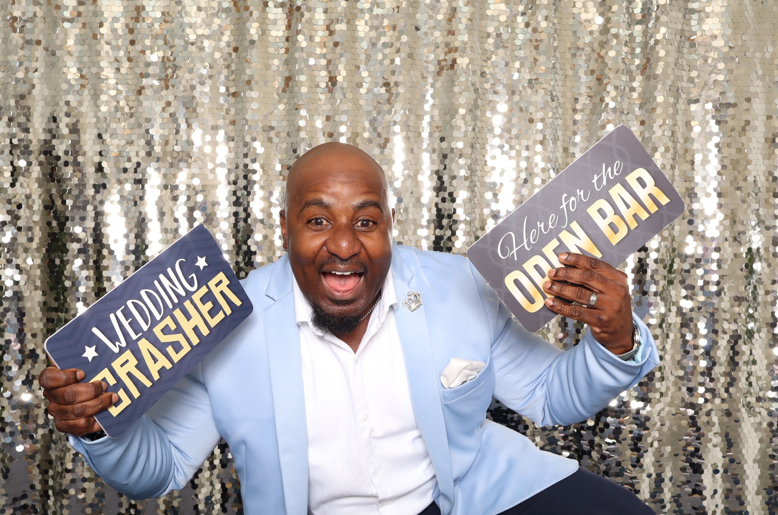 Man in blue suit holding Wedding Crasher and Open Bar signs at a glittery photo booth backdrop.