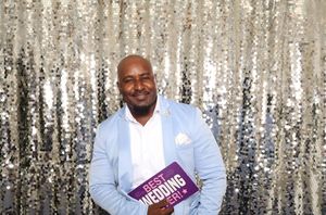 Man in light blue blazer holding a Best Wedding award, smiling in front of a silver sequin backdrop.