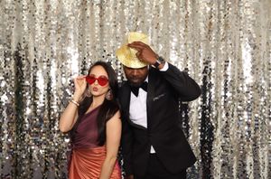 Two people posing at a photo booth with sequin backdrop, woman in red sunglasses and man in gold sequin hat and tuxedo.