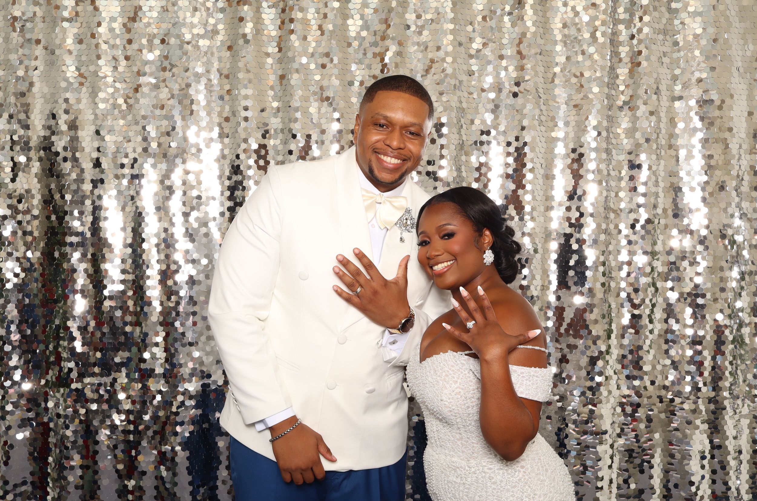 A joyful couple in formal wedding attire poses in front of a silver sequin backdrop, showing off their rings.