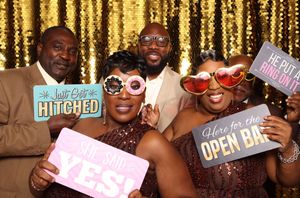 Five friends pose joyfully at a wedding photo booth with fun signs including Hitched, Open Bar, and She Said Yes.