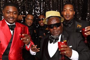 Group of men in formal attire toasting with drinks at an elegant event with a glittery backdrop.