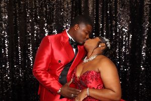 Couple sharing a romantic kiss at formal event, man in red tuxedo and woman in red gown, black sequin backdrop.