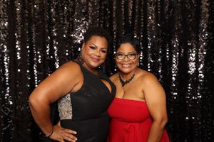 Two women smiling and posing together at formal event wearing elegant dresses against black sequin backdrop.
