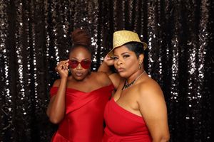 Two women in red dresses pose playfully in front of a black sequin backdrop, one wearing red sunglasses and one a gold hat.