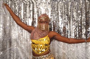 Dancer in gold sequin costume and embellished face veil poses arms wide before shimmering silver sequin backdrop.