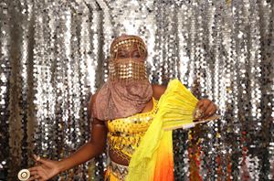 Woman in golden belly dance costume with veil and yellow scarf poses before a silver sequin backdrop.