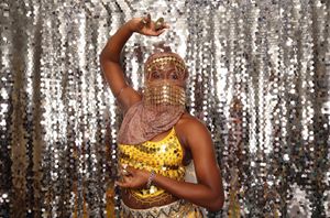 A dancer in gold sequin costume and beaded face veil poses against a shimmering silver sequin backdrop.