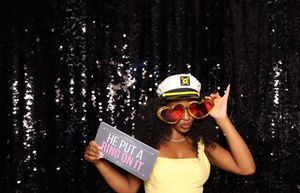 Woman in captain hat and heart sunglasses holds 'He Put a Ring on It' sign at a glittery black photo booth.