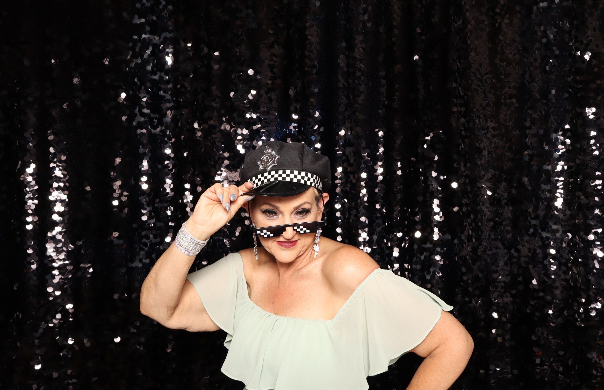 Woman in white dress posing with checkered sunglasses and headband against black sequin backdrop.