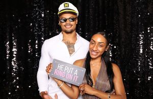 Couple posing at photo booth with sparkly backdrop, man in captain hat holding “He Put a Ring On It” sign.