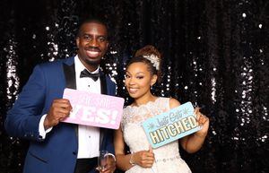 Newlywed couple posing at wedding photo booth holding signs saying YES and Just Got Hitched against black sequin backdrop.