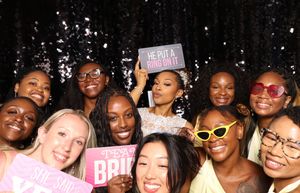 Group of women celebrating a bachelorette party in a photo booth with fun props and signs.