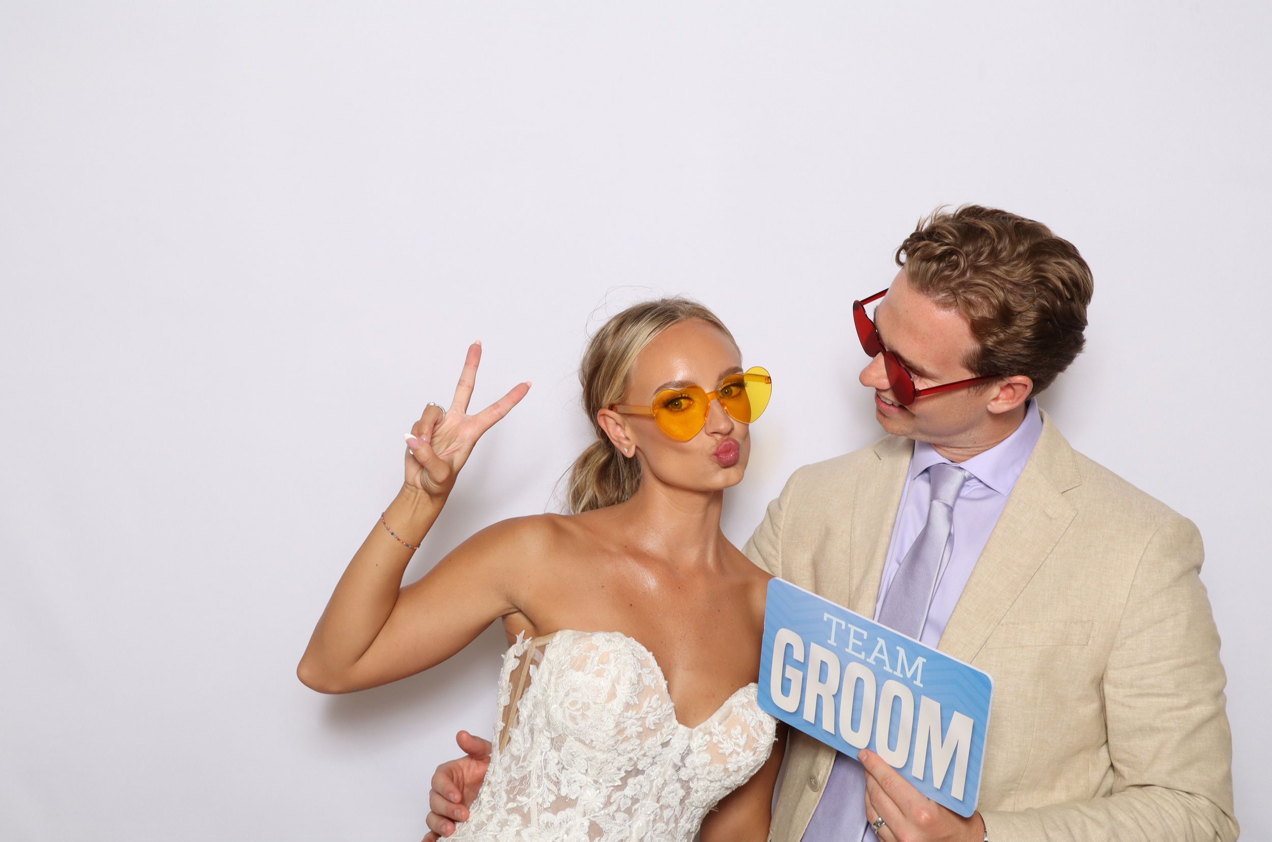 Bride and groom posing in a fun photo booth, wearing colorful sunglasses and holding a Team Groom sign.