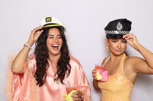 Two women laughing in a photo booth wearing captain and police hats, holding drinks in festive outfits.