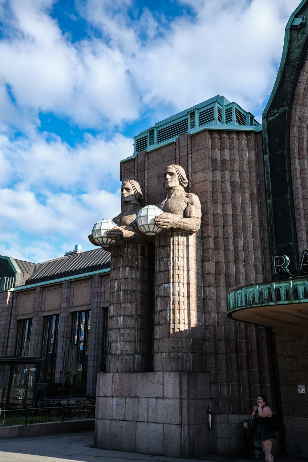Stone statues holding globe lanterns flank the entrance of Helsinki Central Station under a blue cloudy sky.