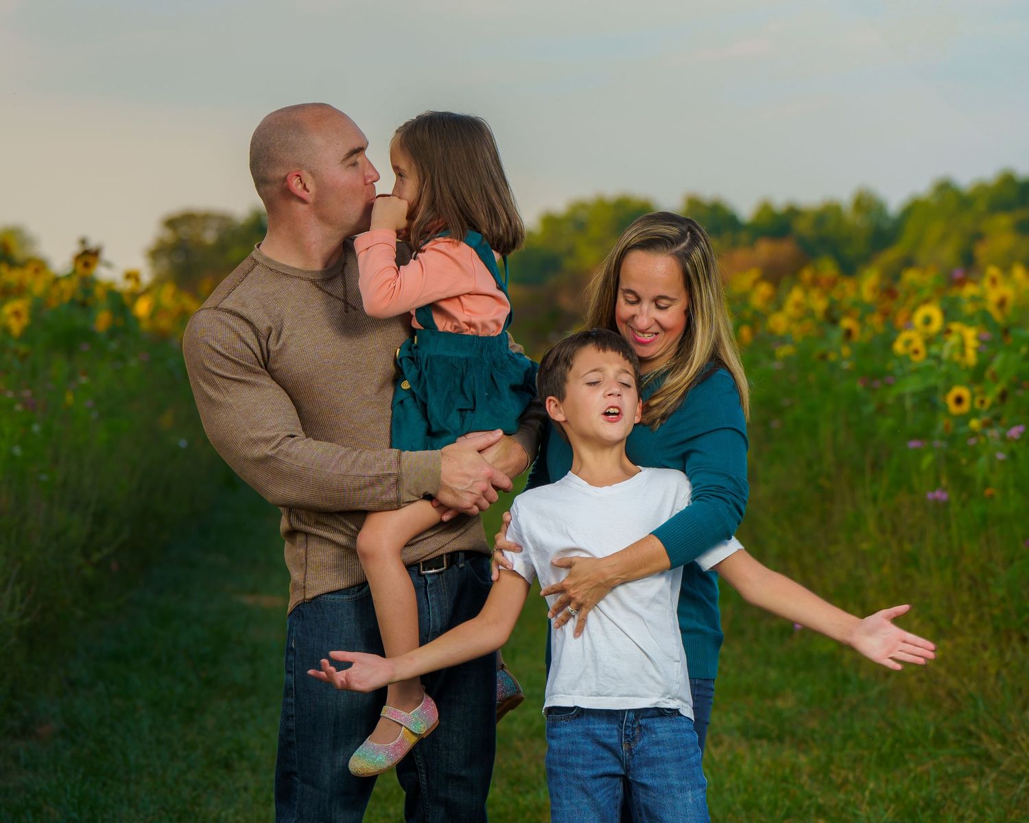 Family of four posing in sunflower field, parents holding children, smiling outdoors at golden hour.