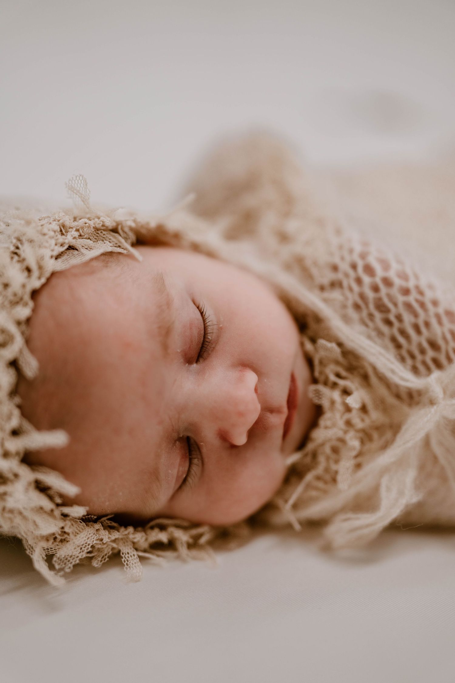 Sleeping newborn baby wrapped in a delicate cream lace bonnet and knit wrap on a white background.
