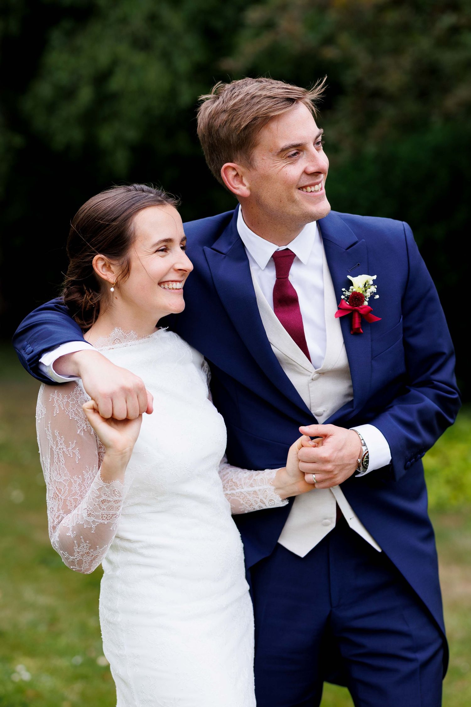 A smiling bride and groom in wedding attire embrace outdoors, with the groom in a navy suit and burgundy tie.