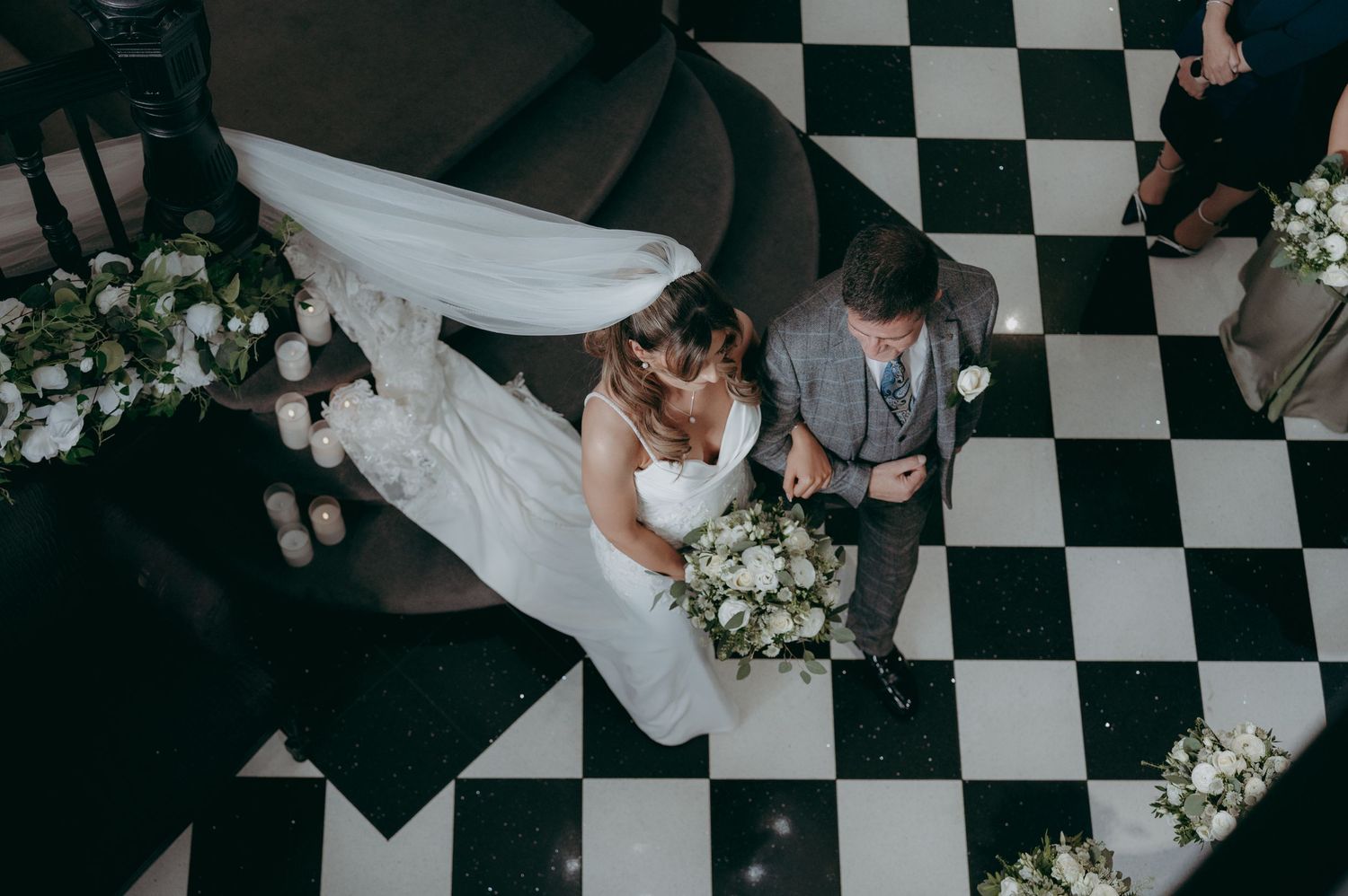 Aerial view of bride and groom walking on black and white checkered floor surrounded by white floral arrangements.