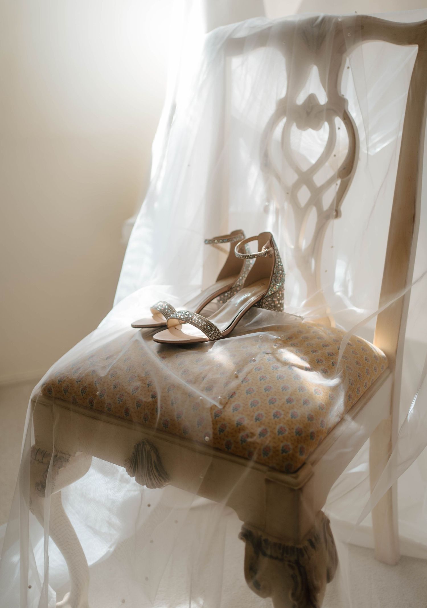 Elegant bridal heels with crystal embellishments resting on a vintage chair draped with sheer white fabric.