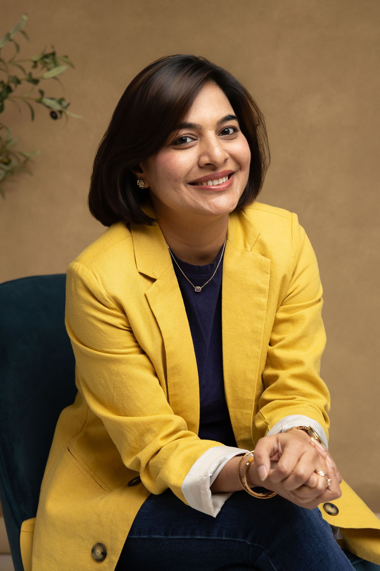 Woman in yellow blazer smiling, seated confidently with hands clasped, wearing gold jewelry and jeans.