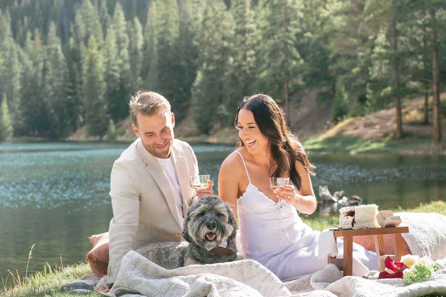 Couple in wedding attire sharing wine with their dog by a mountain lake on a picnic blanket during a Colorado elopement