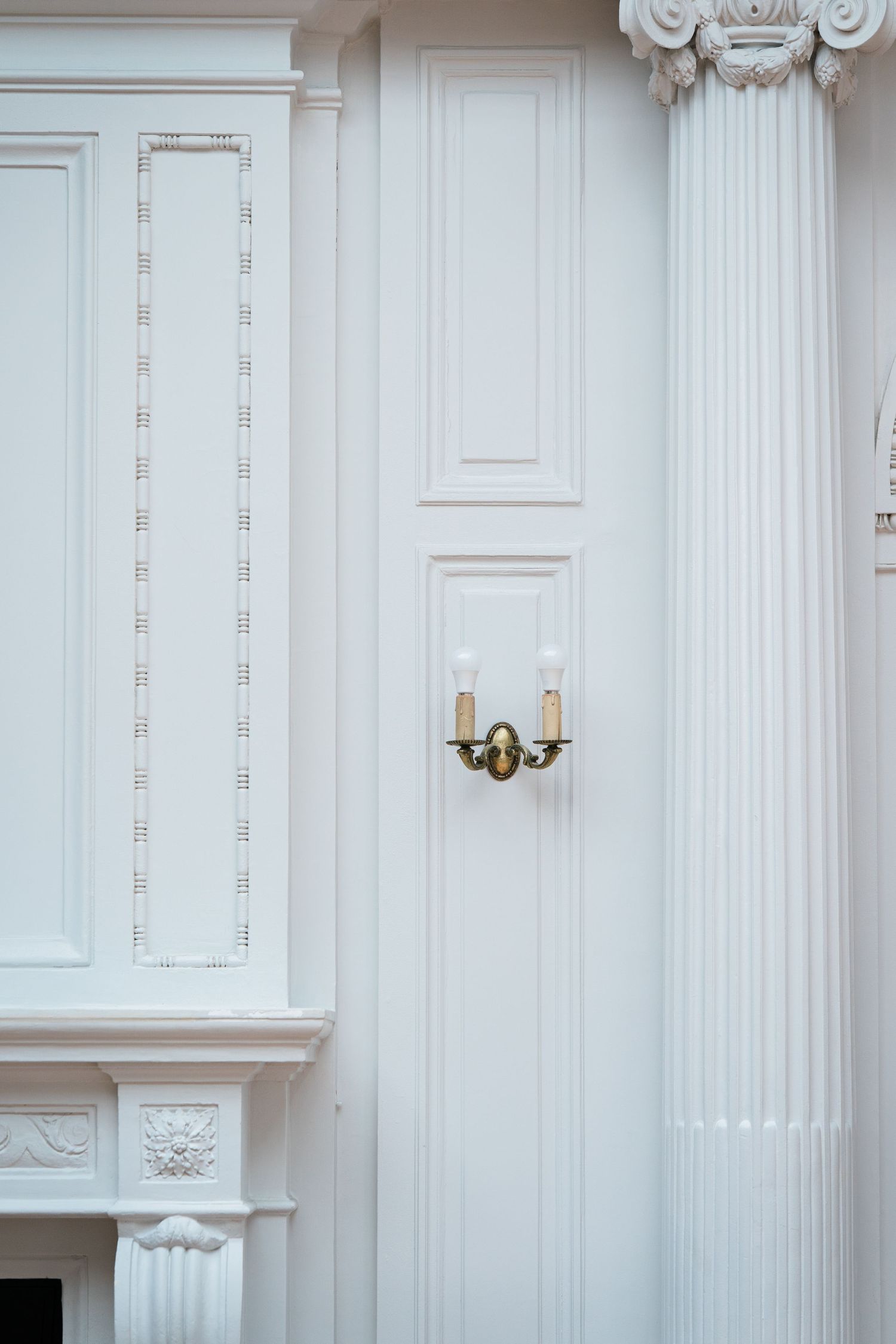 Elegant white interior wall with classical pilaster, ornate moldings, and a gold two-arm wall sconce light fixture.