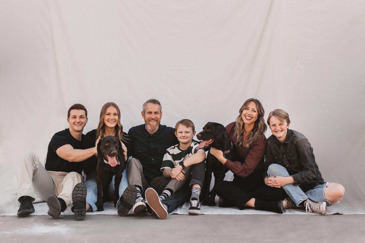 Family of five with two black dogs posing together, smiling in a casual studio portrait against a white backdrop.