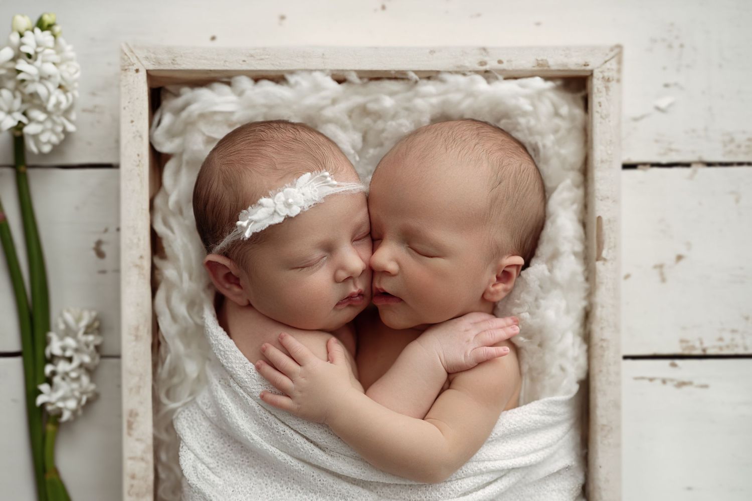 Two sleeping newborn twins wrapped in white, nestled together in a wooden box with soft white filler and flowers nearby.