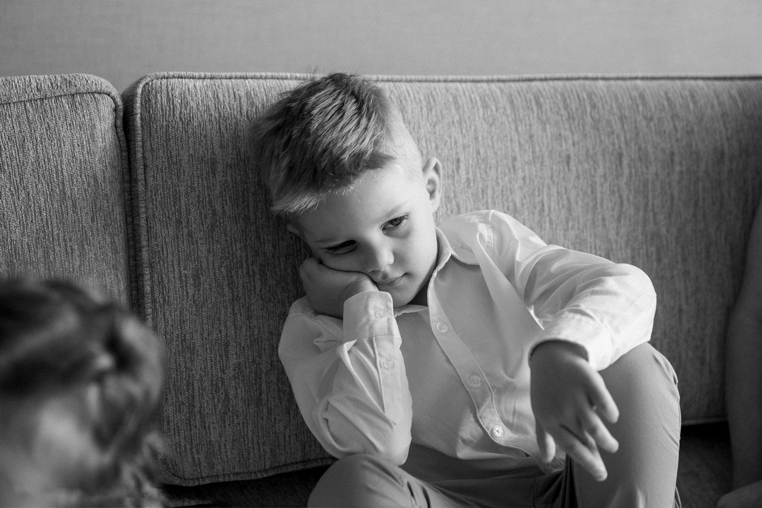 Black and white photo of a young boy in a white shirt sitting on a couch, looking thoughtful and slightly sad.