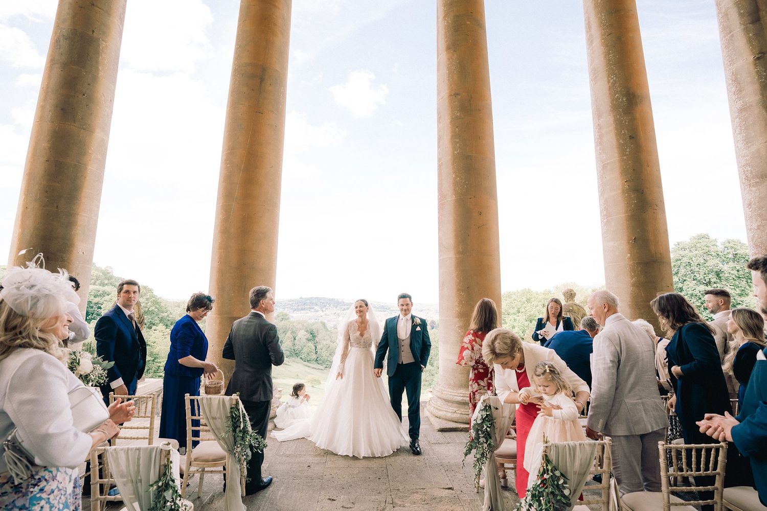 Bride and groom walk down the aisle between tall stone columns at an elegant outdoor wedding ceremony.