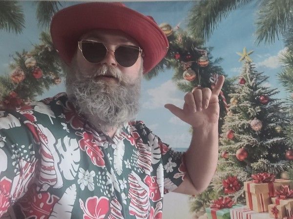 Bearded man in red cap, sunglasses, and tropical Hawaiian shirt poses near a decorated Christmas tree.