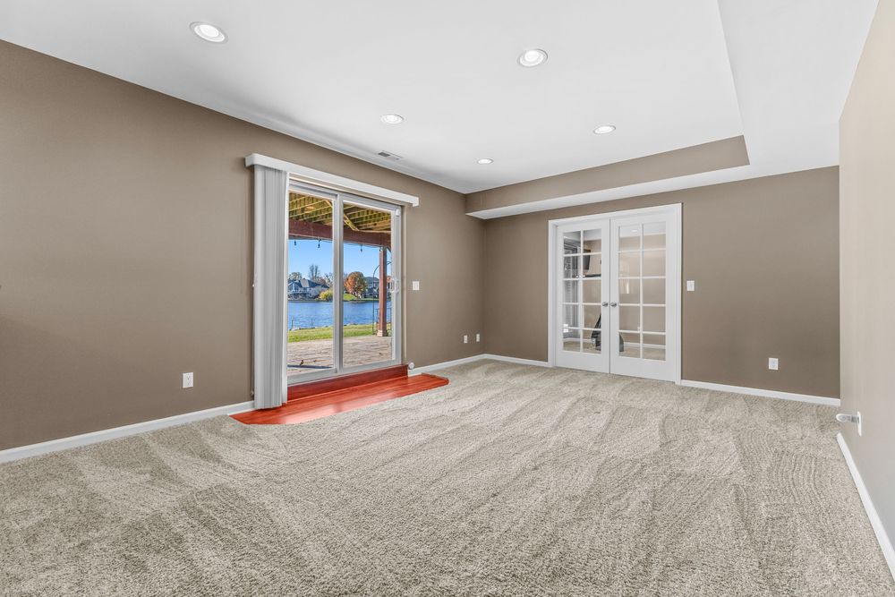 Spacious empty room with beige carpet, tray ceiling, recessed lighting, French doors, and sliding glass doors with waterfront views.