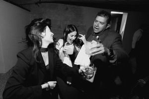 Black and white photo of three young people laughing and sharing drinks at a casual social gathering.