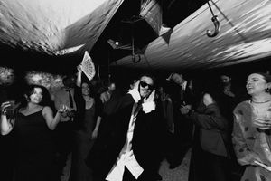 Black and white photo of guests dancing at a formal party event, with a man in sunglasses and suit in the foreground.
