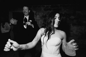 Black and white photo of a bride dancing joyfully at her wedding reception while a man claps behind her.
