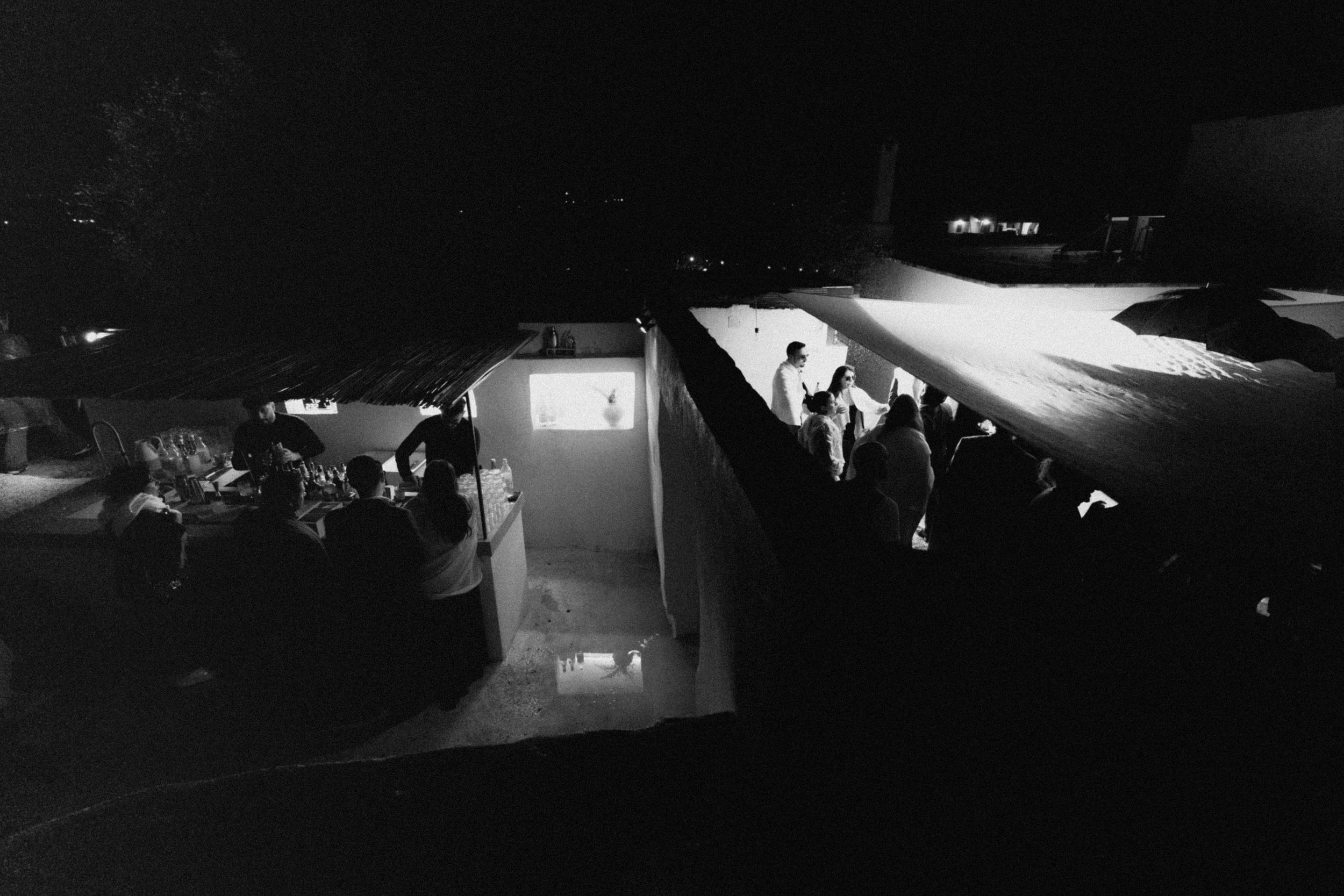 Black and white aerial night photo of a modernist building with illuminated pool and guests gathered on terraces.