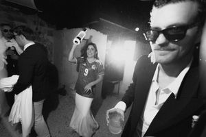 Black and white photo of wedding guests, woman in soccer jersey, man in suit with sunglasses at outdoor reception.