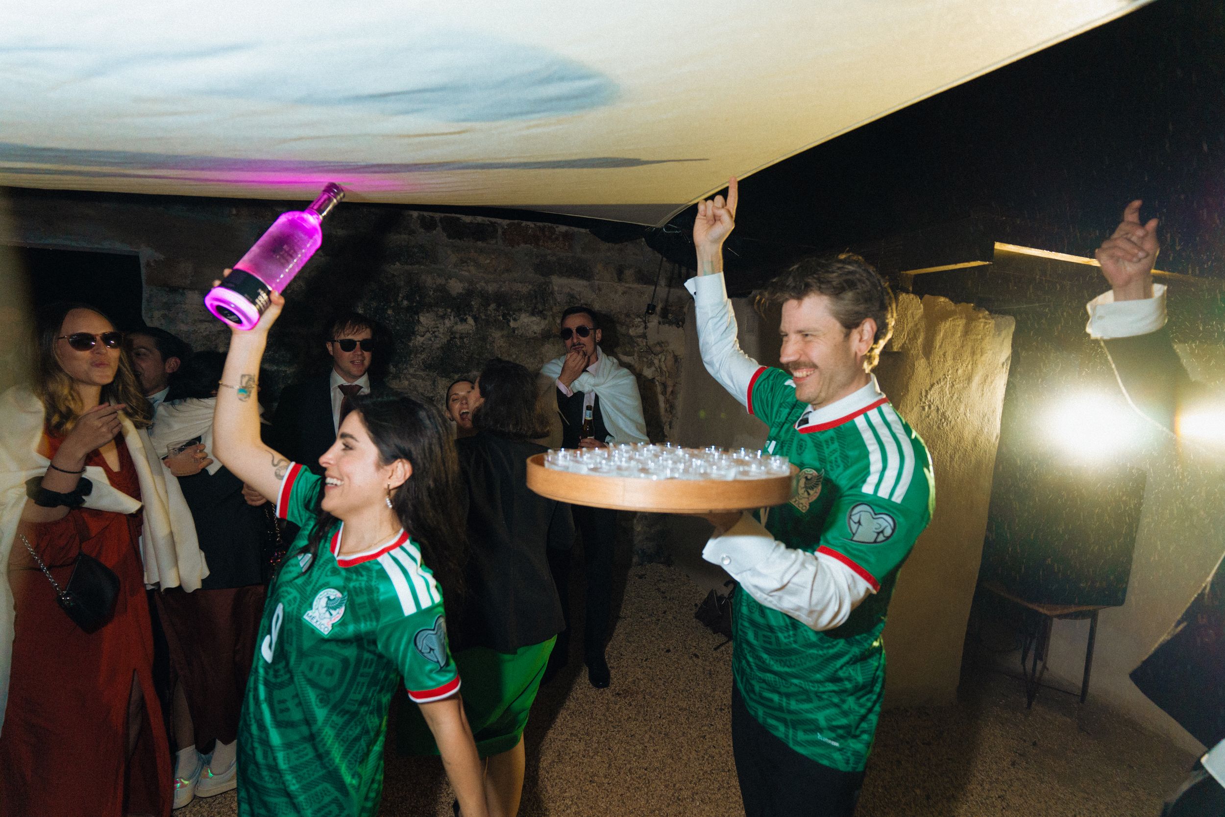 Two people in Mexico soccer jerseys celebrate with a glowing pink bottle and tray of drinks at a lively party.