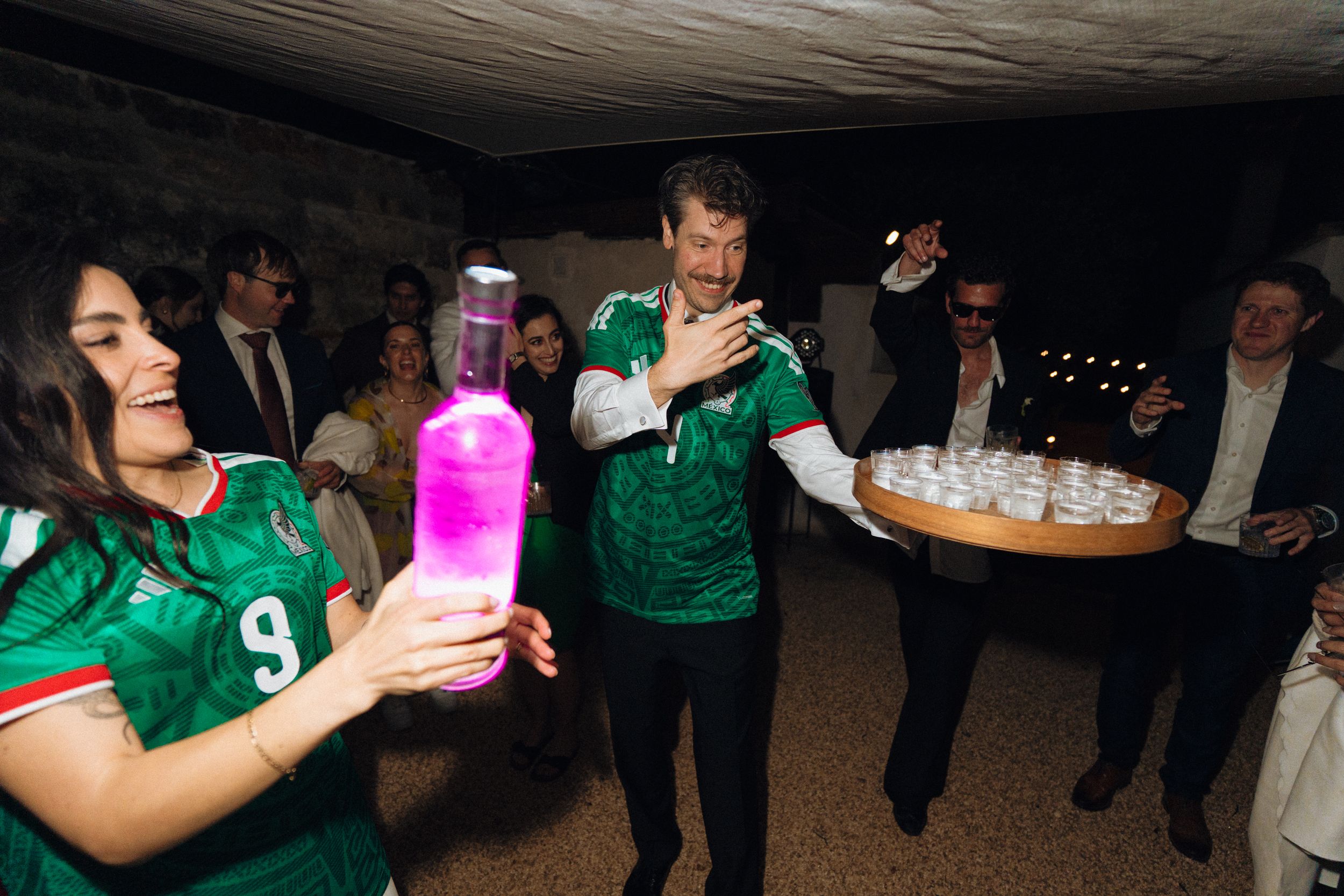 Woman and man in green Mexico jerseys serve drinks with a glowing pink bottle at an indoor celebration.